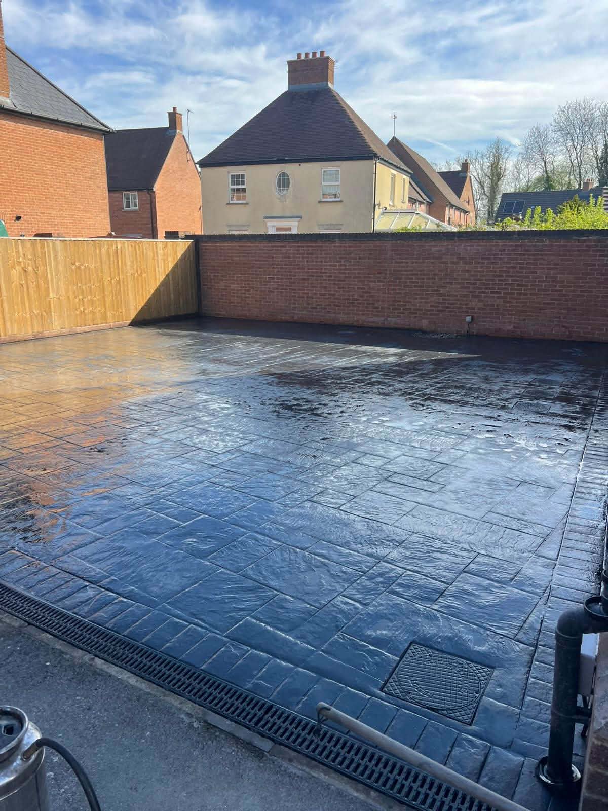 Glossy dark grey pattern-imprinted concrete patio with an integrated drainage channel and manhole cover, enclosed by a brick wall and wooden garden fence.