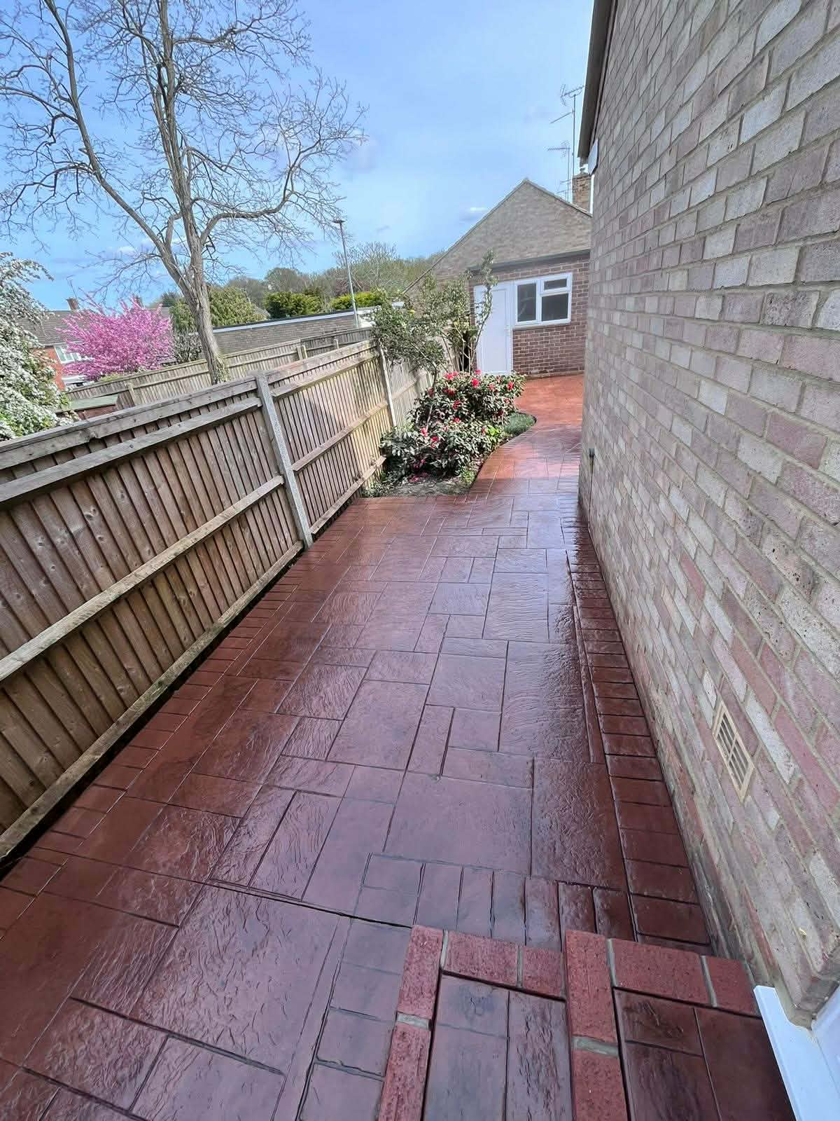 Red-brown pattern-imprinted concrete side pathway and patio area with a textured slate design installed between a brick house and a wooden garden fence.