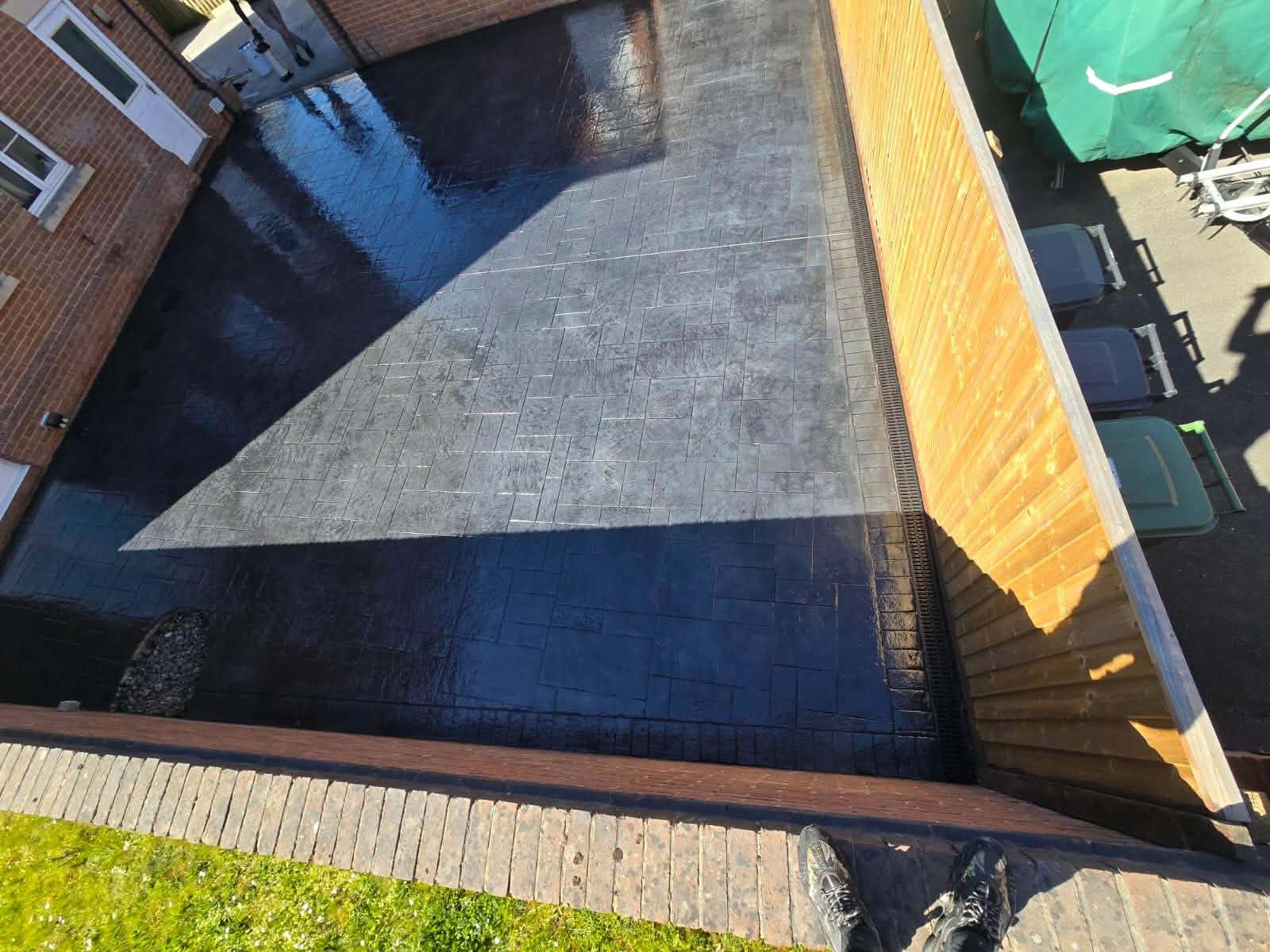 High-gloss dark grey pattern-imprinted concrete patio with a textured stone design and integrated side drainage channel installed behind a brick building.