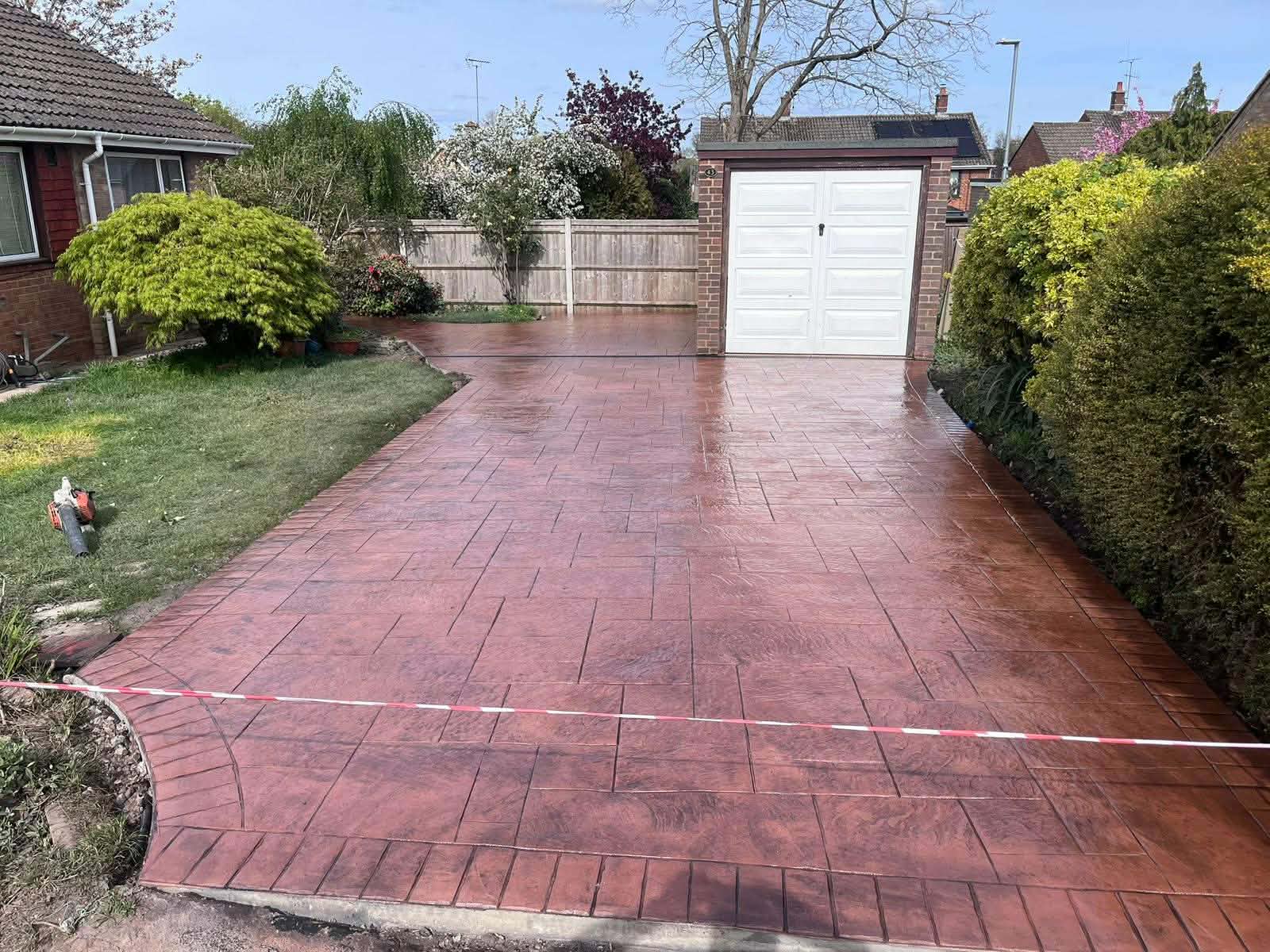 Red-brown pattern-imprinted concrete driveway with a structured tile design and custom border leading up to a detached brick garage with a white door.