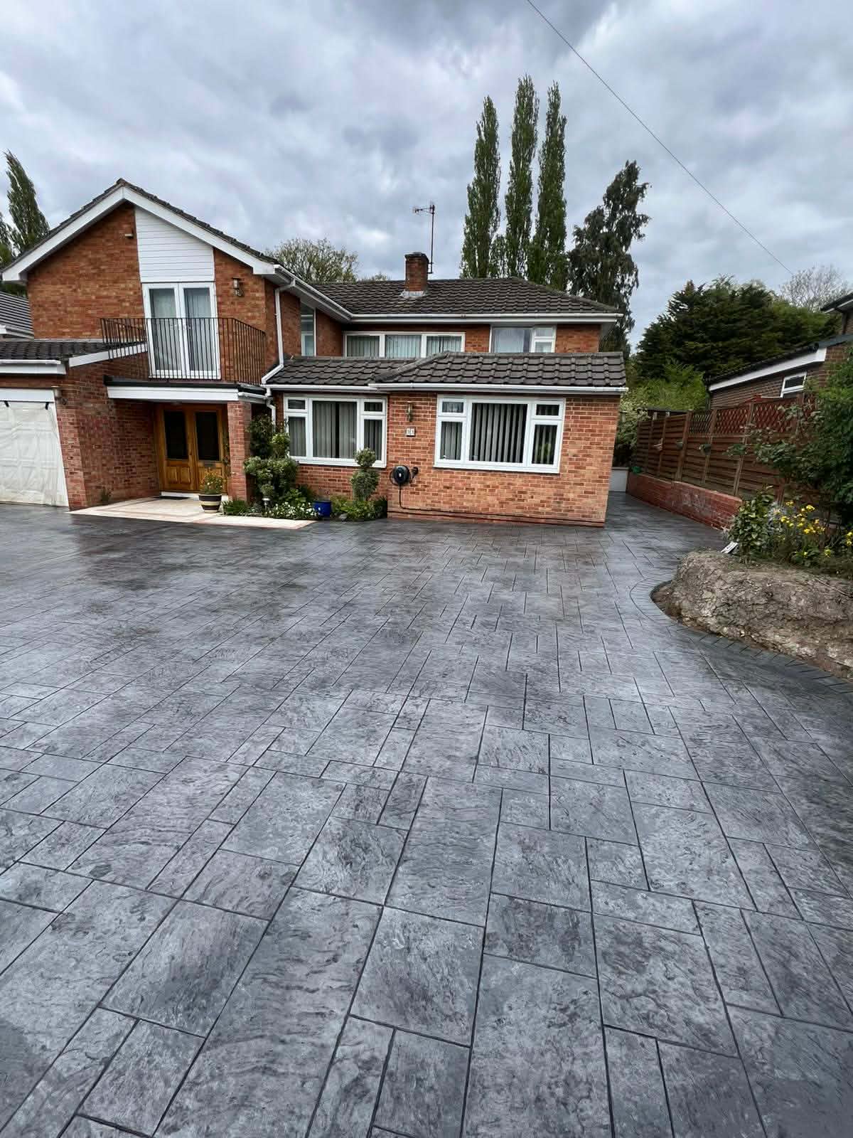 Spacious dark grey textured imprinted concrete driveway installation featuring a stone pattern in front of a large two-storey brick house.