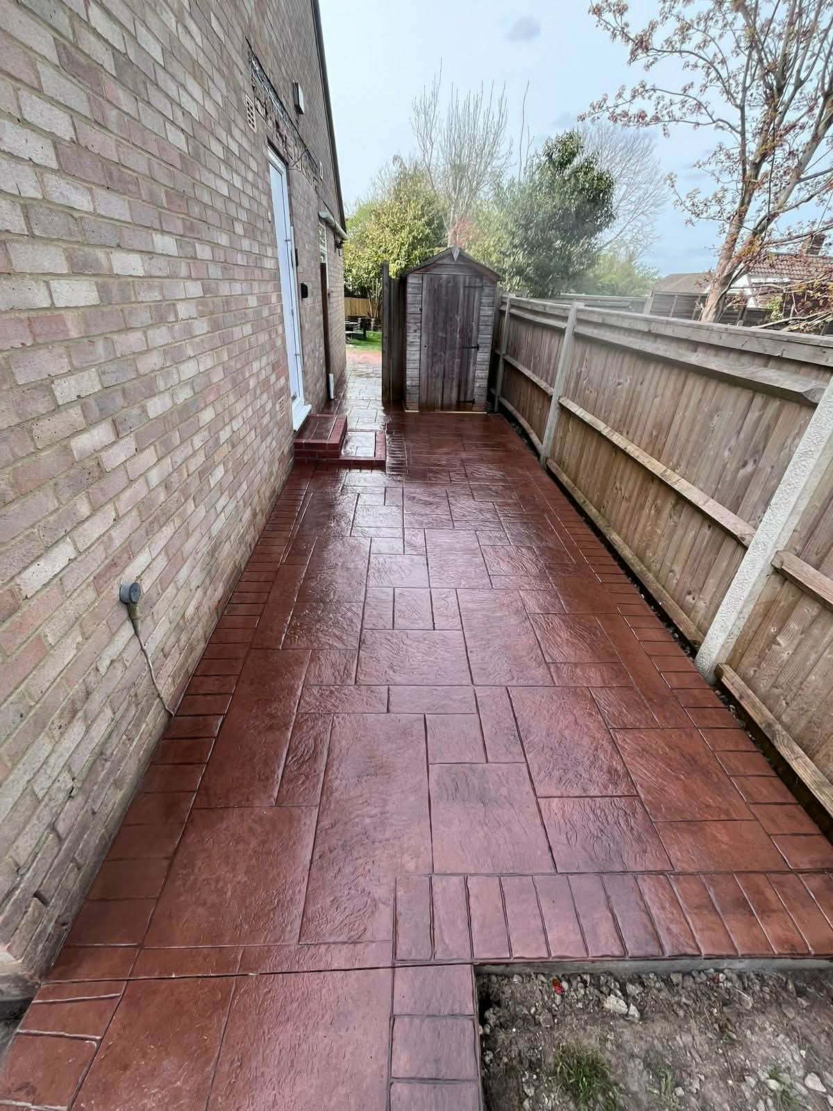 Red-brown pattern-imprinted concrete side pathway and patio area with a textured slate design installed between a brick house and a wooden garden fence.