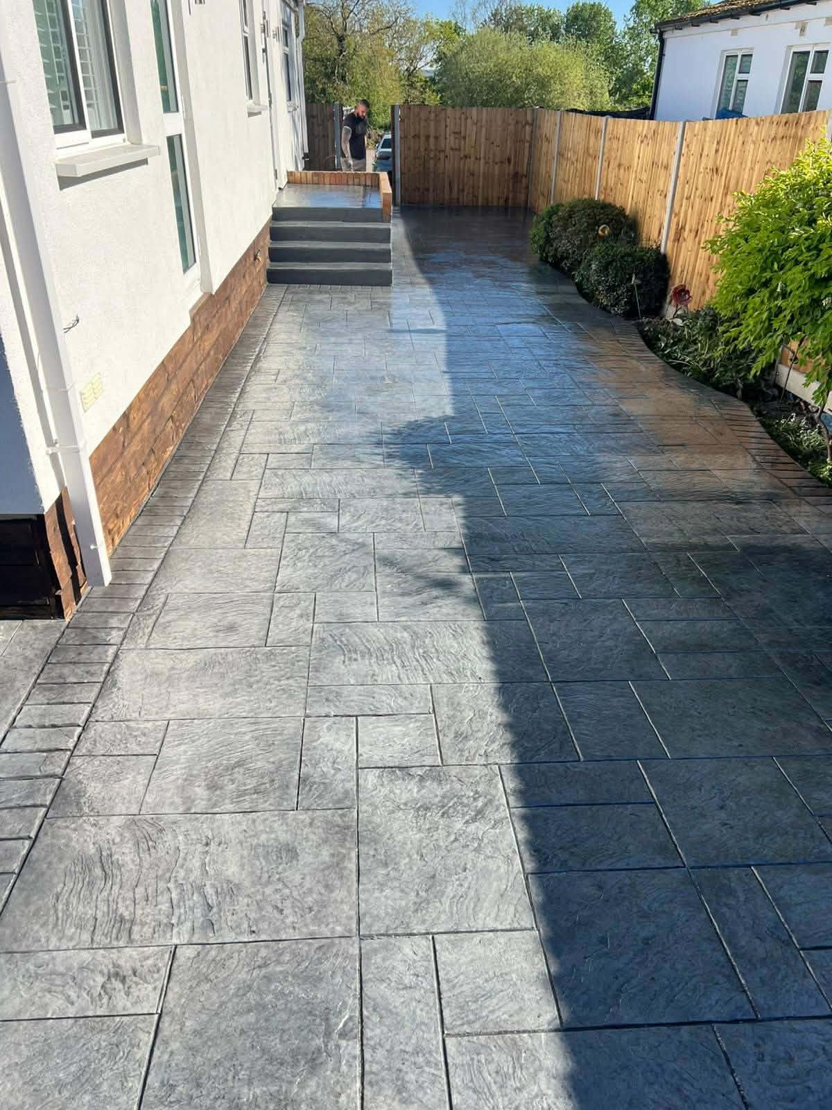 Long textured dark grey pattern-imprinted concrete side driveway and patio area with concrete steps leading up to the entrance of a white residential building.