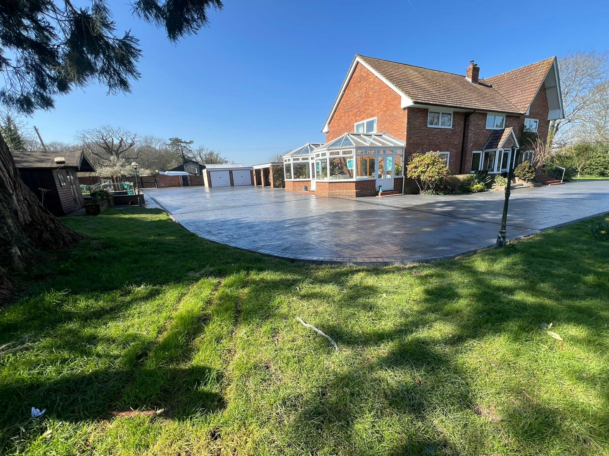 Spacious dark grey imprinted concrete patio and driveway wrap-around installation for a large brick house with two glass conservatories and a green lawn.
