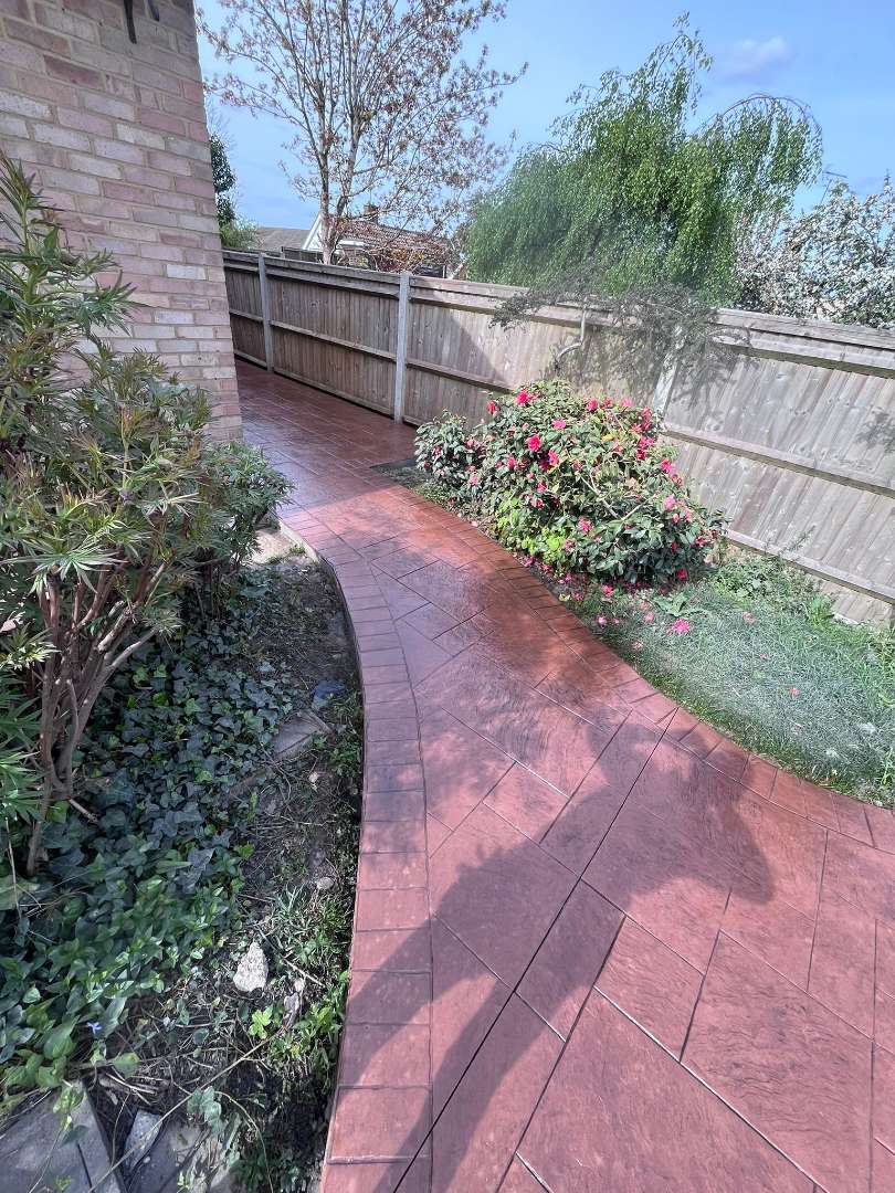A curved, reddish-brown pattern-imprinted concrete garden pathway featuring an ashlar stone design and a structured brick-effect border, running along a brick house wall and a wooden panel fence with green shrubs.