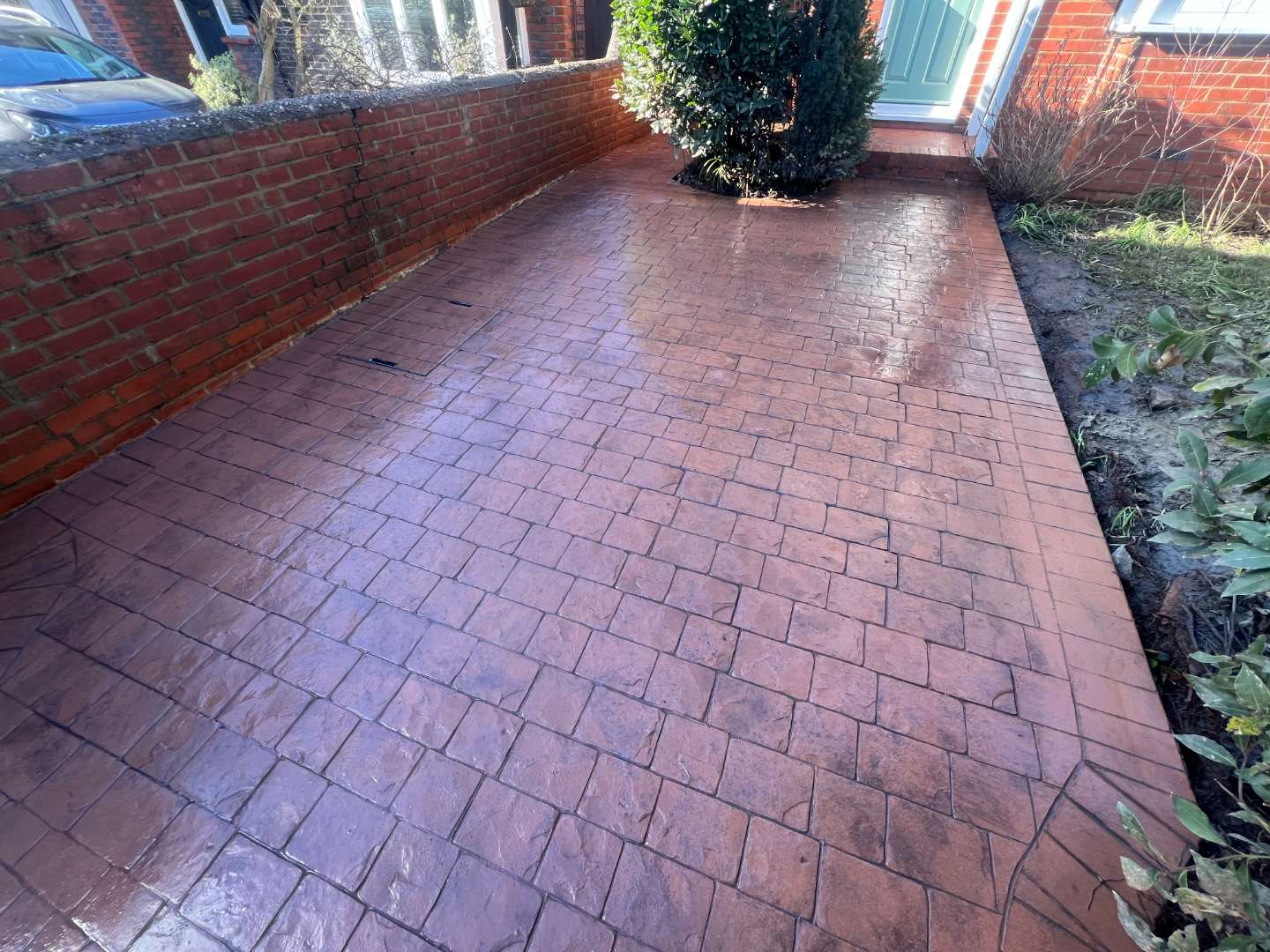 A glossy, reddish-brown pattern-imprinted concrete driveway with a textured block paving design, enclosed by a brick boundary wall on one side and leading to a residential front door with a small green bush.