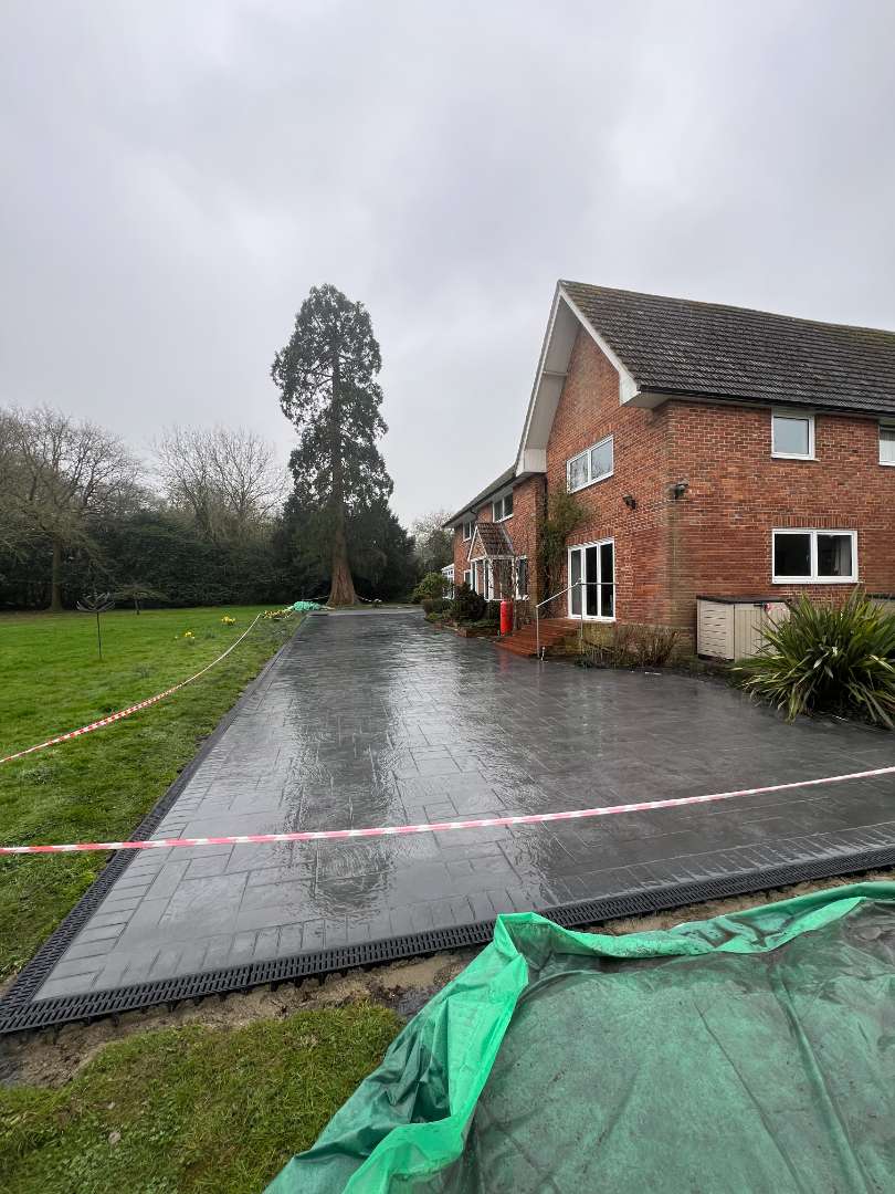 A long, wet dark grey pattern-imprinted concrete driveway with a structured brick-effect border and integrated drainage channels, running past a large brick residential house and an expansive green lawn cordoned off with red and white tape.
