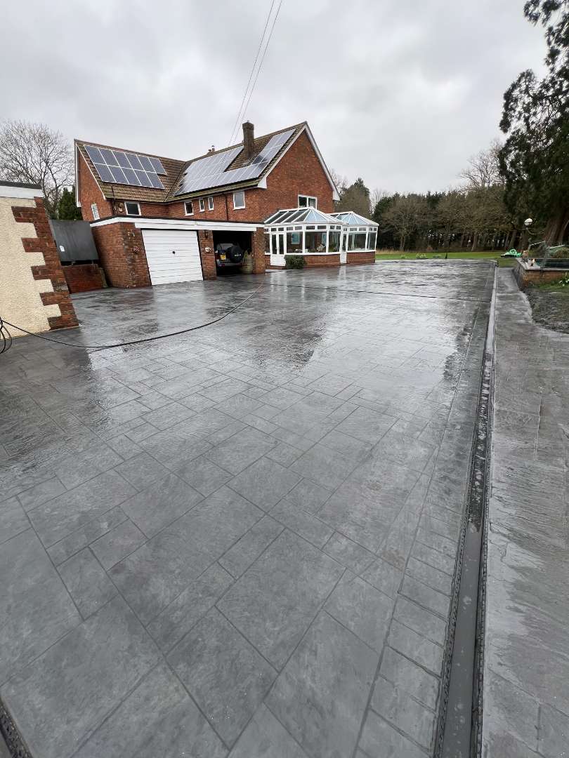 A wide, wet dark grey pattern-imprinted concrete driveway featuring a textured stone tile design and an integrated drainage channel, in front of a brick house with solar panels, an attached garage, and a white conservatory.