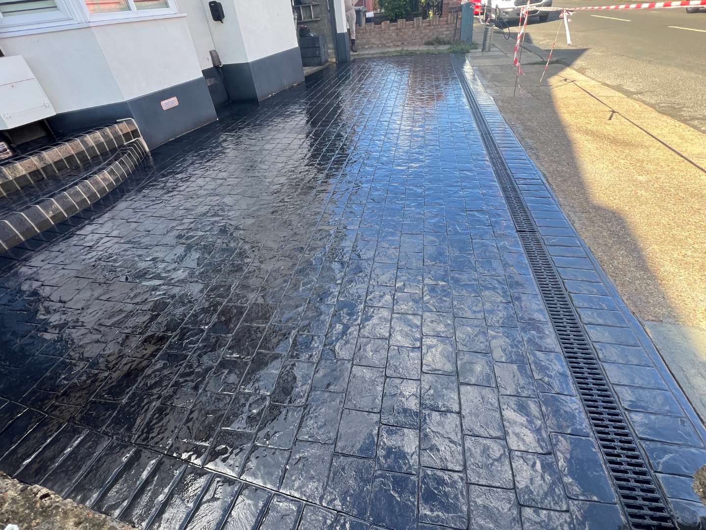 Glossy dark grey pattern-imprinted concrete driveway featuring a cobblestone style design with a long integrated drainage channel running along the edge next to a public pavement.