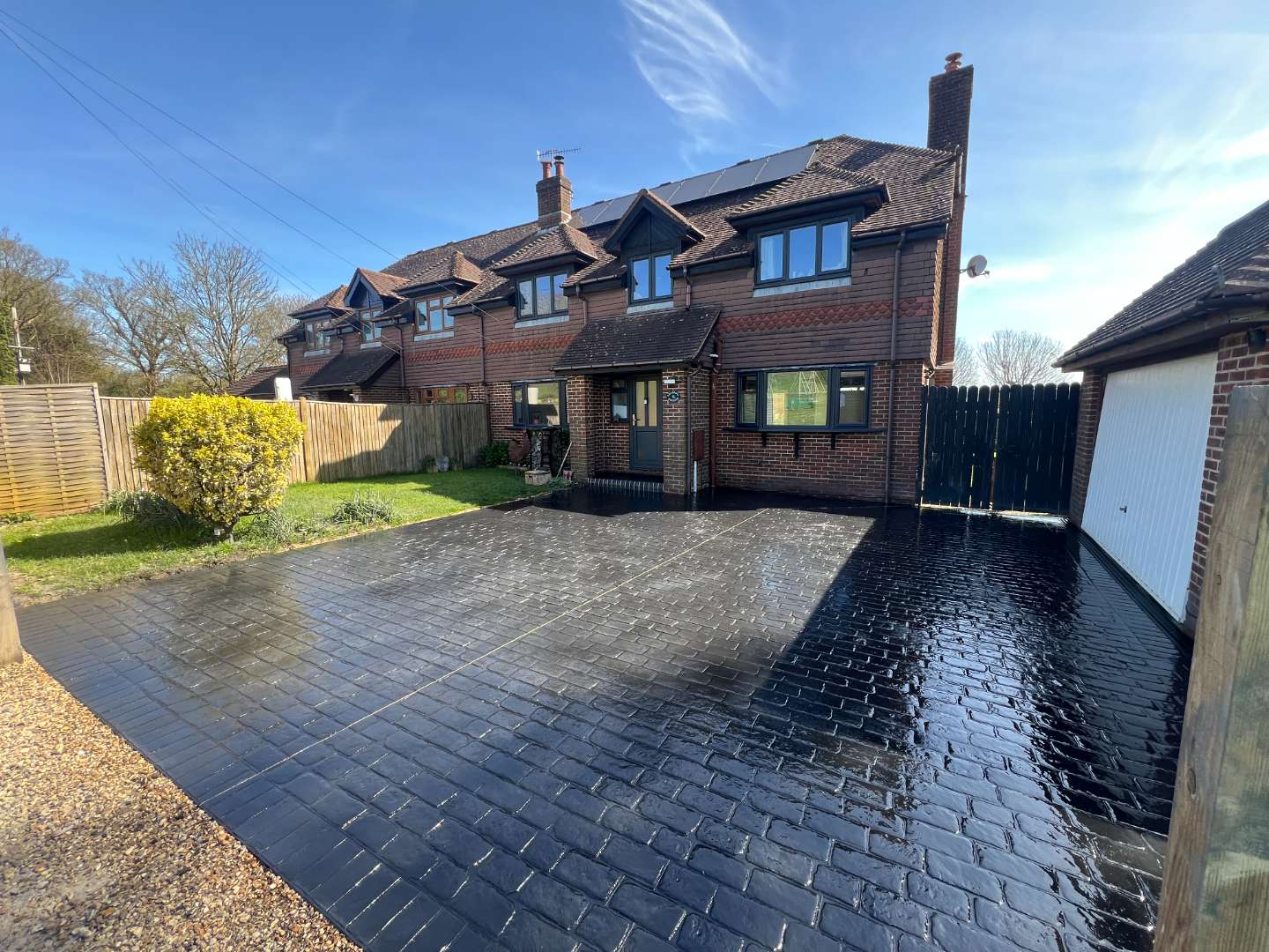 Glossy dark grey pattern-imprinted concrete driveway featuring a cobblestone style design in front of a multi-storey brick semi-detached house on a sunny day.