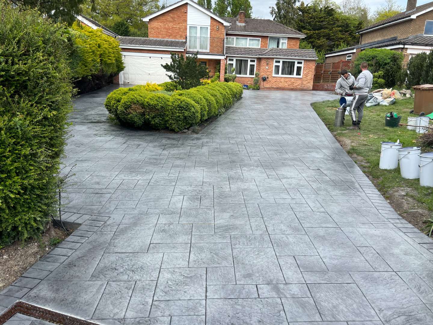 Two contractors working on a wide dark grey pattern-imprinted concrete driveway with a textured ashlar slate design, next to a landscaped central garden bed in front of a brick house.