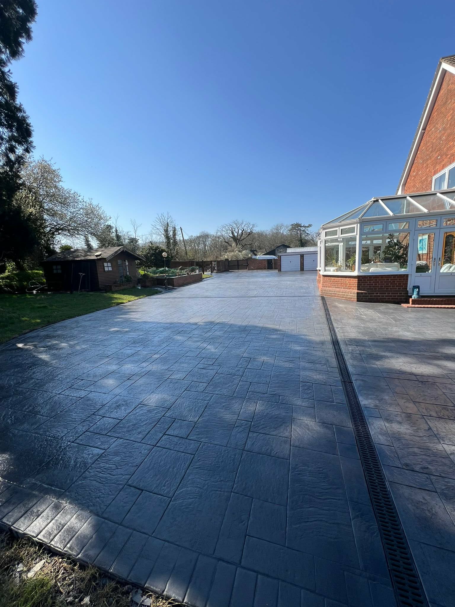 Long pattern-imprinted concrete driveway in dark grey featuring an integrated drainage channel next to a brick home with a white conservatory.