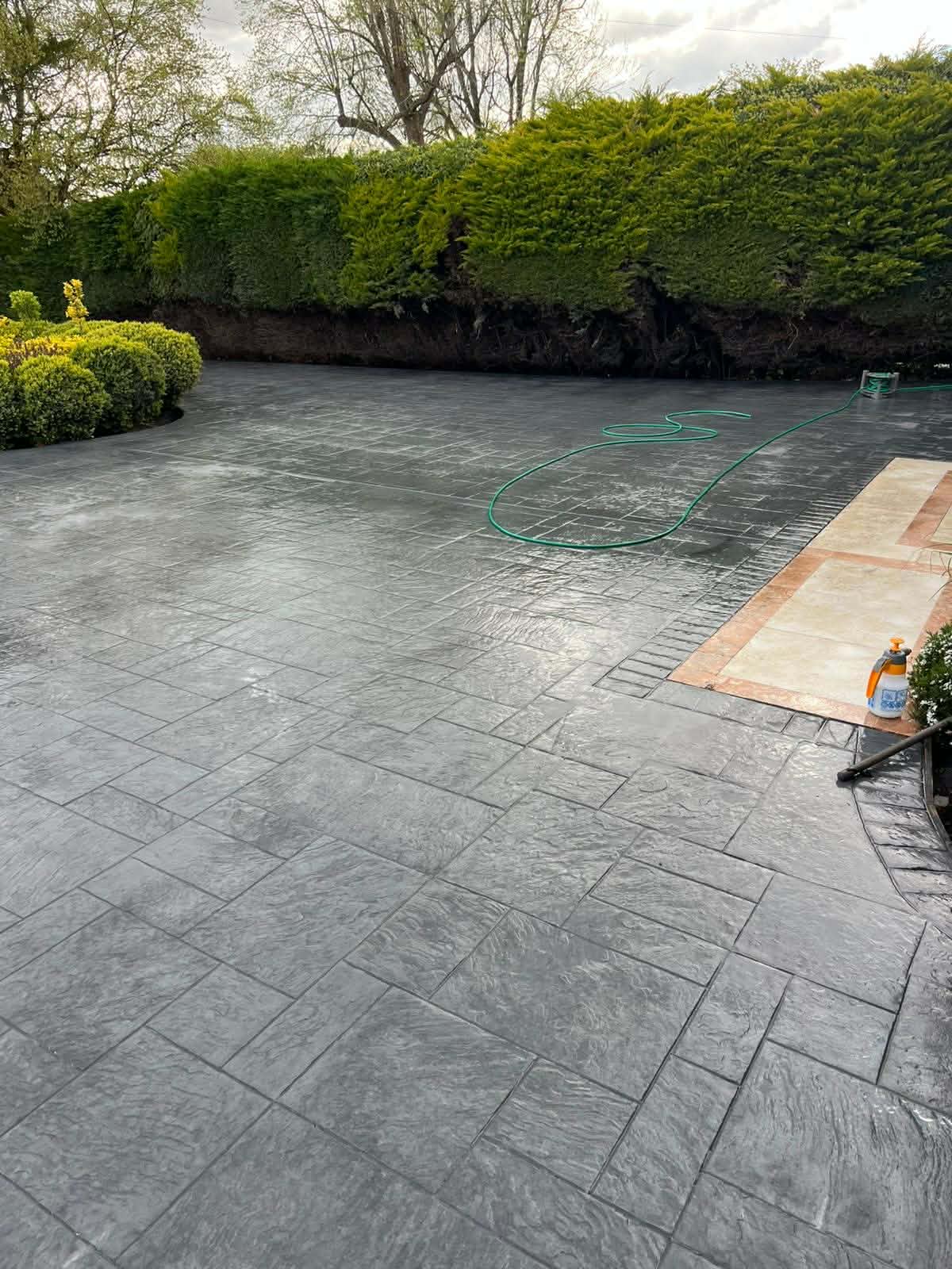 Large dark grey textured imprinted concrete driveway and patio installation featuring a stone pattern and custom borders next to high green garden hedges.