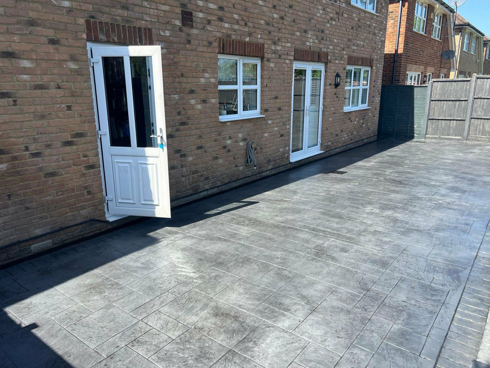 Newly installed dark grey pattern-imprinted concrete patio with a textured ashlar slate design running along the back of a brick house with white double doors.