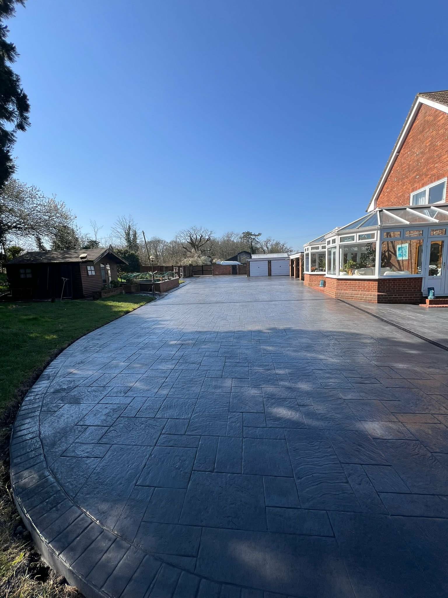 Large, newly installed dark grey imprinted concrete driveway and patio area alongside a brick house with a conservatory by VVMS Driveways in London.