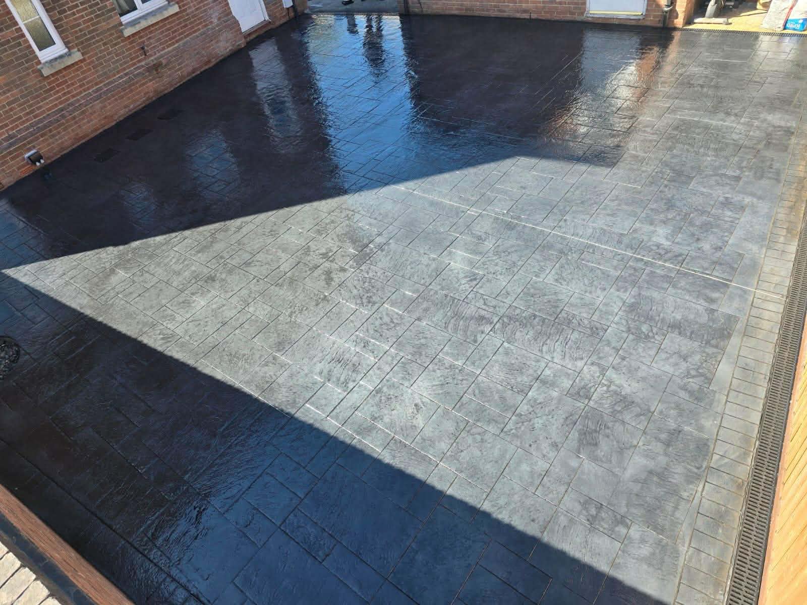 High-gloss dark grey pattern-imprinted concrete patio with a textured stone design and integrated side drainage channel installed behind a brick building.