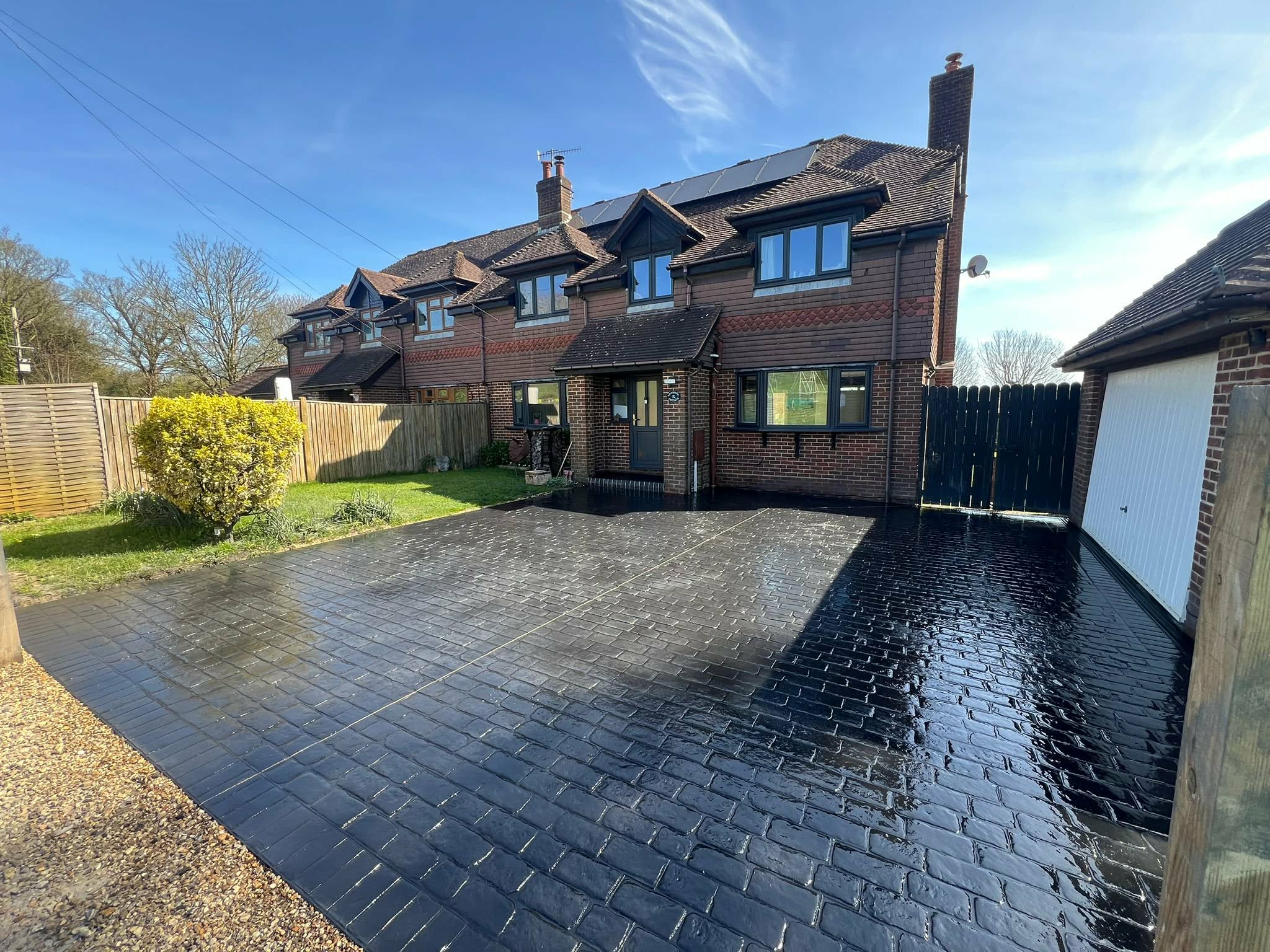 Glossy black cobblestone-style imprinted concrete driveway and front parking area for a brick semi-detached house with a detached garage under a clear blue sky.